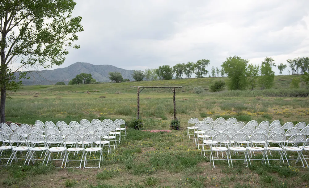 A rustic timber wedding arch standing in an open meadow at Bear Creek Lake Park in Lakewood, white folding chairs set in rows with foothills behind