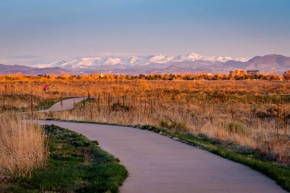 A paved trail winding through open grassland at Cherry Creek State Park in Aurora, with the snowcapped Front Range lit at golden hour