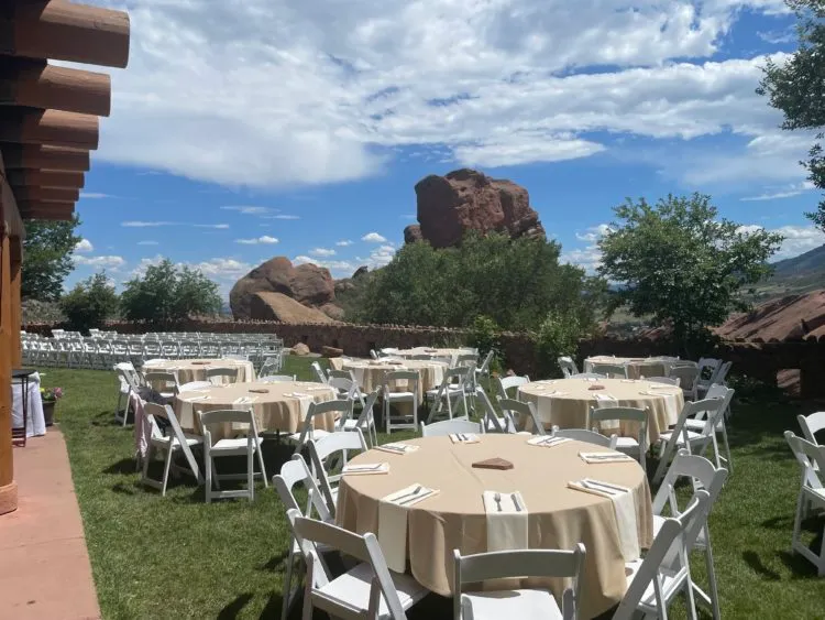 Round reception tables with tan linens on a lawn at Red Rocks Park in Morrison, red sandstone formations rising behind