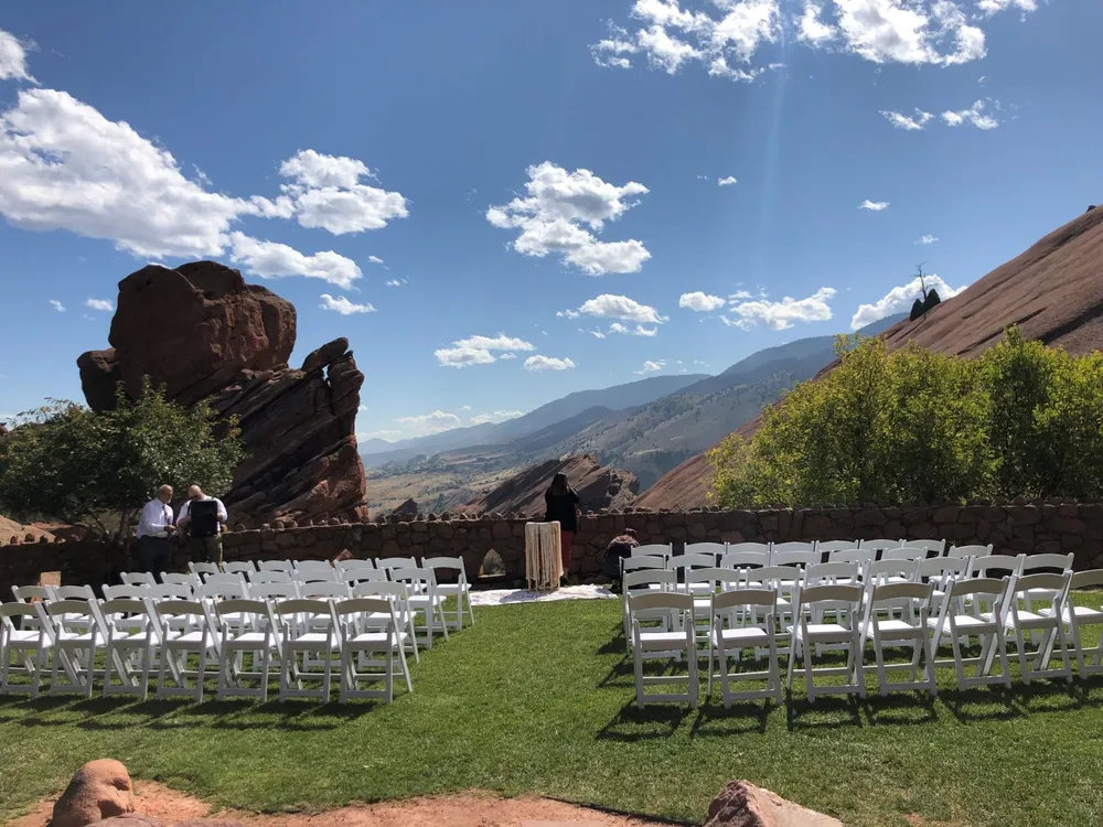 Ceremony chairs arranged on a lawn at Red Rocks Park in Morrison, facing a stone wall with red rock formations and mountain views beyond