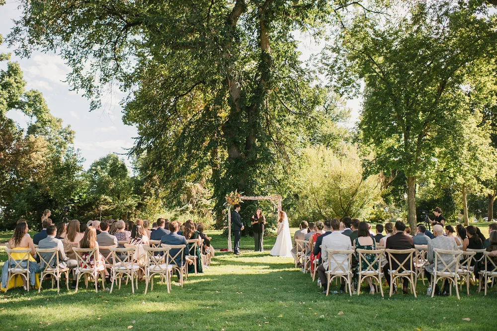 A wedding ceremony beneath mature trees at Washington Park in Denver, guests seated on wooden cross-back chairs facing a floral-topped wooden arch